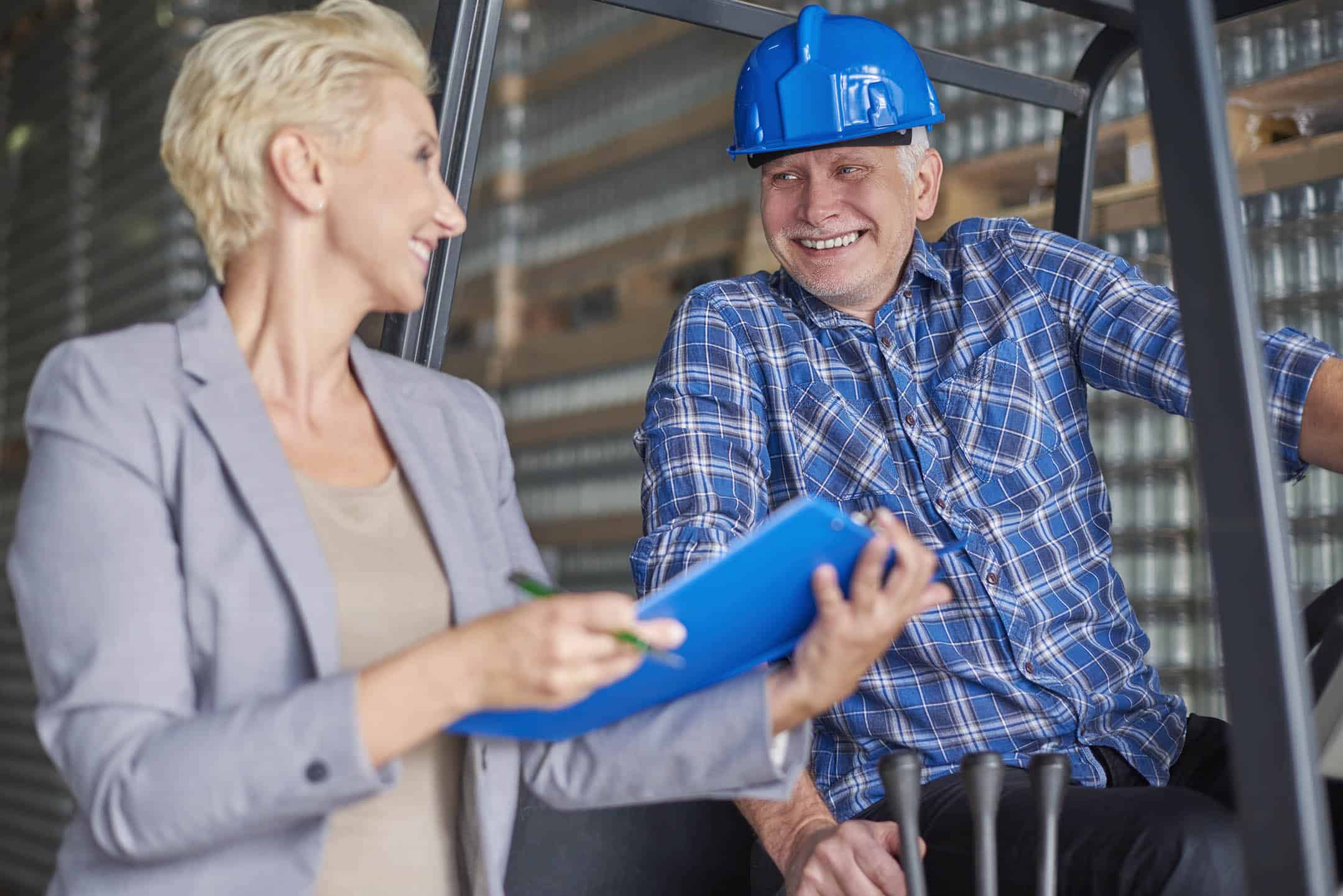 Tradesman on a ladder smiling at a smart business woman holding a clipboard.